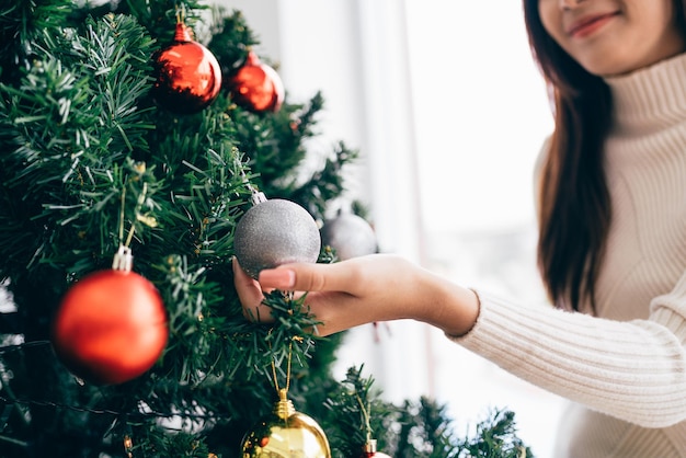 Feche a mão da mulher tocando e decorando uma bugiganga vermelha na árvore de Natal na sala de estar em casa Desfrutando e celebrando o feriado de Natal