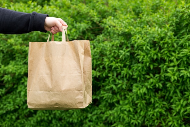 Feche a mão do homem com um saco de papel para comida para viagem em fundo verde da natureza. entrega em qualquer tempo ao redor do relógio para o cliente.