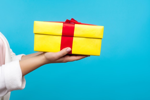 Feche a mão feminina no suéter branco segurando a caixa de presente embrulhada amarela dando presente no aniversário ou dia de boxe celebração do feriado de natal caridade estúdio tiro isolado no fundo azul