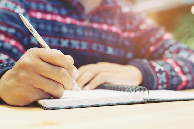 Feche a mão jovem está sentado usando a caneta para escrever o bloco de notas de aula de registro no livro sobre a madeira da mesa.