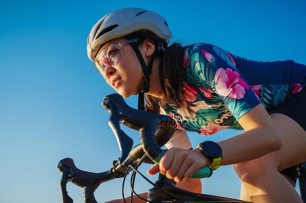 Feche a mulher asiática com forma de corpo atlético em capacete protetor e óculos andando de bicicleta no céu azul