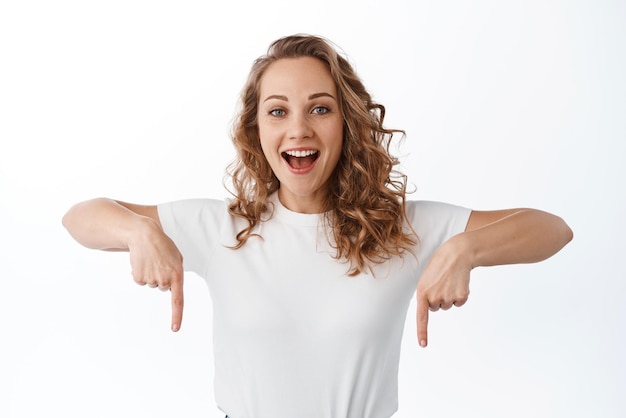 Feche a mulher loira feliz apontando os dedos para baixo em seu logotipo mostrando propaganda e sorrindo em pé contra o fundo branco