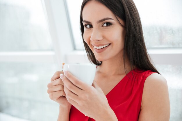 Feche a mulher sorridente com uma xícara de café em pé perto da janela no escritório e olhando para a câmera