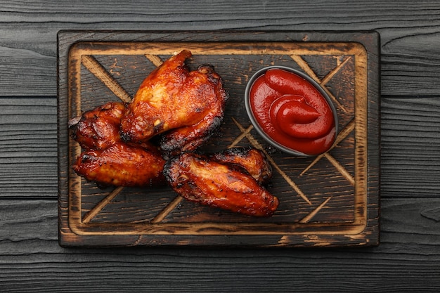 Feche a porção de asas de frango de churrasco grelhado e molho de tomate para churrasco servido na tábua de madeira sobre pranchas de mesa preta, vista superior elevada, diretamente acima