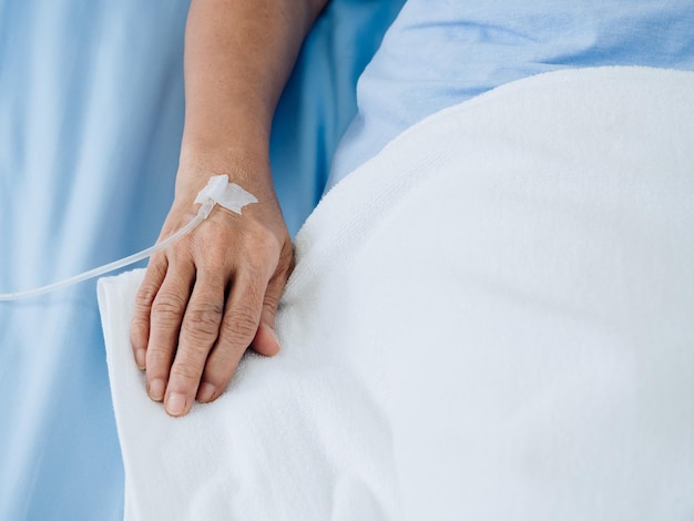Feche a solução salina na mão de pacientes idosos no vestido azul, deitada na cama com cobertor branco no quarto do hospital com espaço de cópia Mão do paciente idoso recebendo solução salina intravenosa