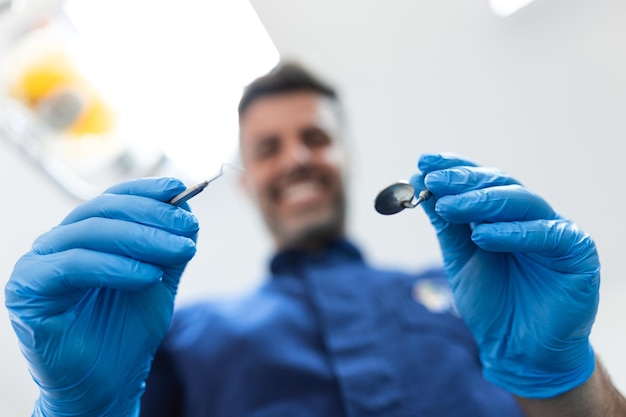 Feche a visão dos pacientes de um dentista Bonito dentista em luvas cirúrgicas está segurando ferramentas e olhando para a câmera
