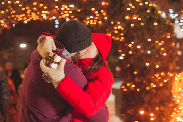 Feche a vista do jovem casal beijando ao ar livre à noite. Mulher segurando uma pequena caixa de presente. Luzes de Natal no fundo. Conceito de estilo de vida feliz