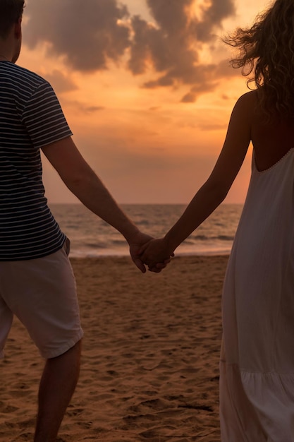 Feche a vista traseira jovem adorável casal de família de mãos dadas homem e mulher em roupas casuais caminhando juntos na praia arenosa no fundo do pôr do sol do oceano ao ar livre