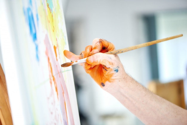 Feche acima da mão masculina do artista, guardando a escova e pintando a pintura alaranjada rica no estúdio do pintor.