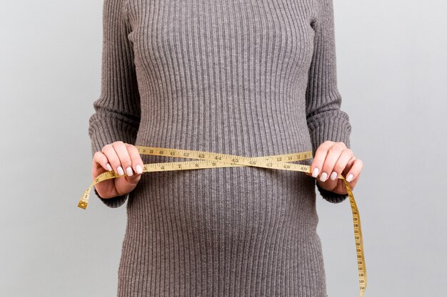 Feche acima da mulher gravida no vestido cinzento que mede sua barriga crescente no fundo cinzento. Esperando um bebê. Medição em polegadas.