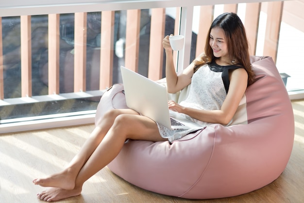 Foto feche acima da vista da mulher, aproveitando o tempo em casa. mulher jovem e bonita sorridente trabalhando no laptop, telefone inteligente e usar fones de ouvido enquanto está sentado em uma cadeira grande e confortável em casa.