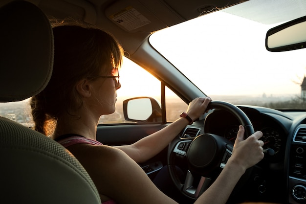 Feche acima da vista de um motorista de mulher segurando o volante dirigindo um carro ao pôr do sol.