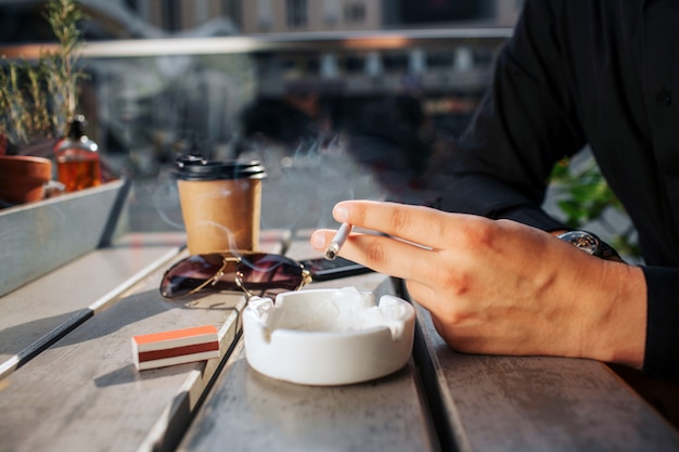 Feche acima das mãos do homem mantenha o cigarro disponivel. Ele está acima do prato branco. Há copos e copos de bebida no talbe. Está ensolarado lá fora.