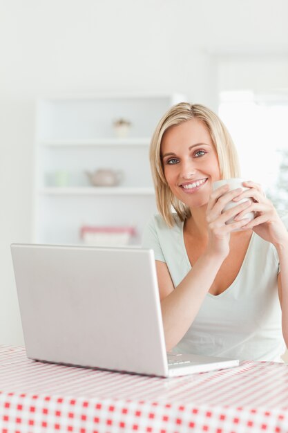 Feche acima de uma mulher segurando xícara de café com laptop na frente dela, sorrindo para a câmera