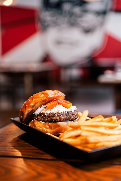 Foto feche acima do cheeseburger com carne grelhada, queijo de feta e o tomate cortado servido com fritadas na placa preta.