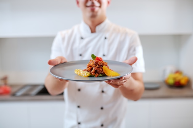 Feche acima do cozinheiro chefe caucasiano no uniforme que guarda a placa com salmão e fruta alaranjada.