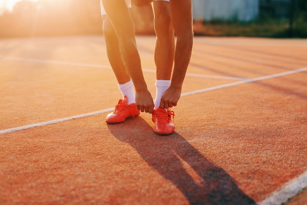 Feche acima do homem desportivo que dobra e que amarra o laço ao estar na corte na manhã no verão.