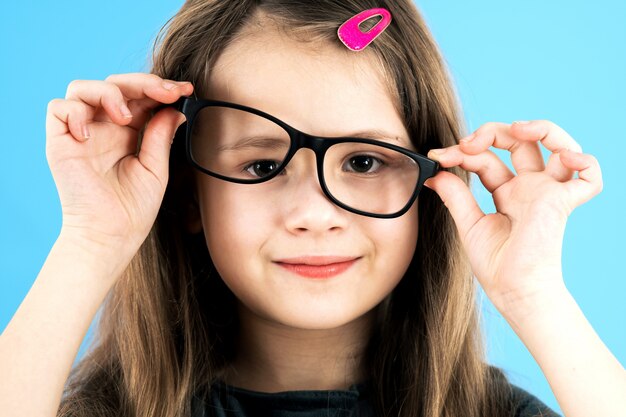 Feche acima do retrato de uma menina engraçada da escola infantil que veste os vidros isolados no fundo azul.