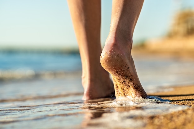 Feche acima dos pés da mulher que andam com os pés descalços na praia da areia na água do mar. conceito de férias, viagens e liberdade. pessoas relaxando no verão.