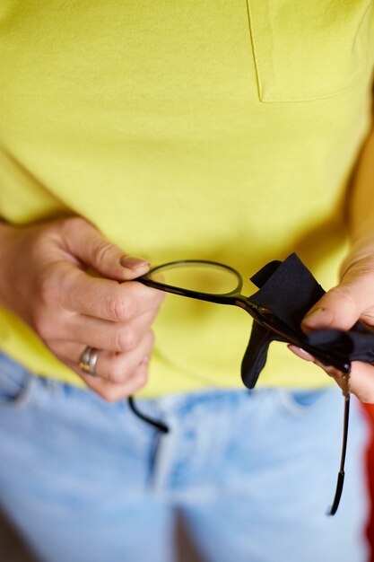 Feche as mãos cortadas de uma mulher limpando lentes de óculos pretos da moda com uma toalha, lenço de microfibra preto