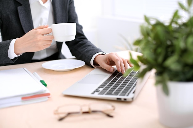 Feche as mãos da mulher de negócios com uma xícara de café está sentado à mesa e digitando em um computador portátil no escritório de cor branca.