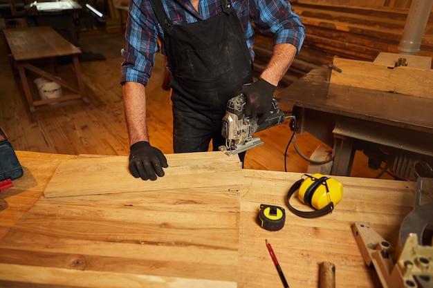 Feche as mãos de carpinteiro profissional serrando madeira com um quebra-cabeça elétrico na oficina de carpintaria
