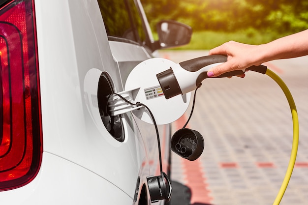 Feche as mãos de mulher, conectando um cabo de alimentação ao carro elétrico para carregar na estação de carregamento ao ar livre.