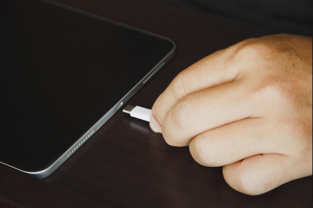 Feche as mãos de um homem usando um cabo USB tye C para carregar um tablet na mesa de madeira