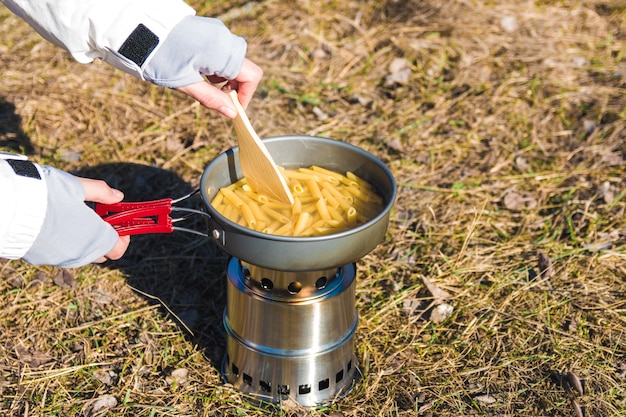 Feche as mãos do alpinista mulher cozinhar macarrão no queimador de madeira em acampamento selvagem