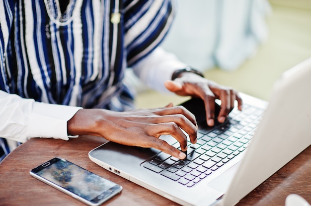 Feche as mãos do homem Africano em roupas tradicionais, sentado atrás do laptop e trabalhando