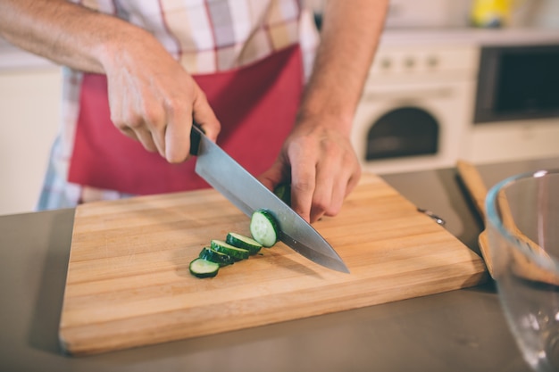 Feche as mãos do homem segurando a faca e corte o pepino em pedaços. ele faz isso na mesa. há uma tigela de vidro na mesa. guy usa pron.