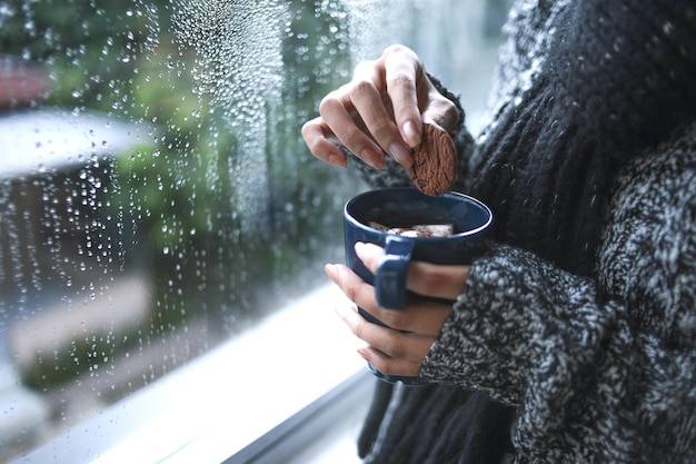 Feche as mãos em malha quente, mergulhando biscoito em café quente perto da janela com gota de chuva