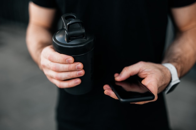 Feche as mãos masculinas de vista de corte usando o smartphone e segurando a garrafa de esportes. tempo de descanso! corredor em camiseta preta. conceito de tecnologias modernas. esporte urbano. plano de fundo cinza.