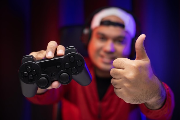 Feche as mãos masculinas segurando o console de jogos de joystick. Jovem jogando videogame online. Homem indiano de jaqueta vermelha joga computador em casa no quarto escuro