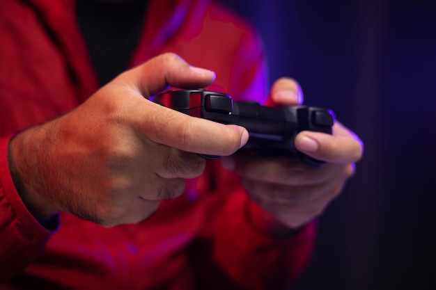 Foto feche as mãos masculinas segurando o console de jogos de joystick. jovem jogando videogame online. homem indiano de jaqueta vermelha joga computador em casa no quarto escuro