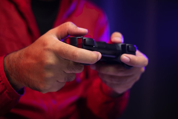 Feche as mãos masculinas segurando o console de jogos de joystick. jovem jogando videogame online. homem indiano de jaqueta vermelha joga computador em casa no quarto escuro