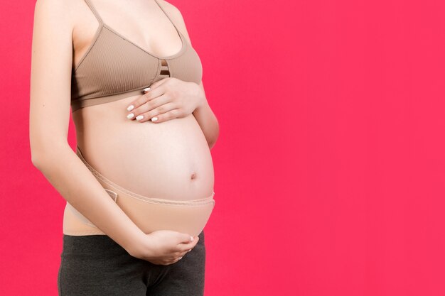 Feche de bandagem de gravidez vestida de mulher grávida para reduzir a dor nas costas em fundo rosa com espaço de cópia. Conceito de cinto de suporte abdominal ortopédico.