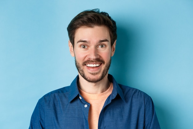 Feche de cara bonito surpreso com barba parecendo divertido, sorrindo feliz para a câmera, de pé sobre fundo azul.