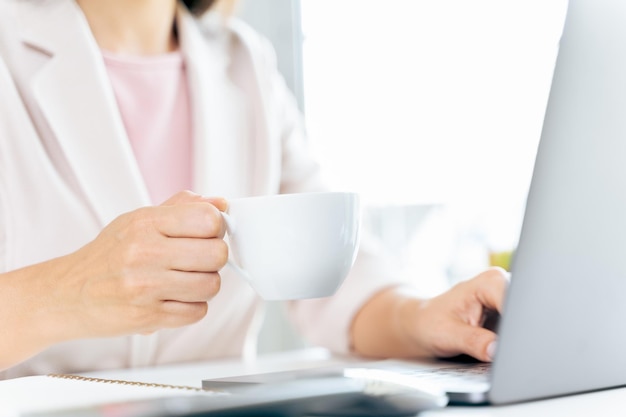 Feche de empresária usando computador portátil e sentado na mesa de escritório, segurando uma xícara de café.