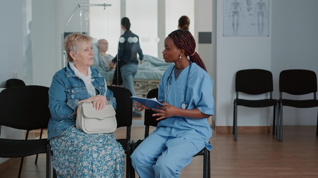 Feche de enfermeira afro-americana falando com o paciente mais velho na área de espera. Assistente médico explicando doença e tratamento de saúde para mulher aposentada na sala de espera na instalação.