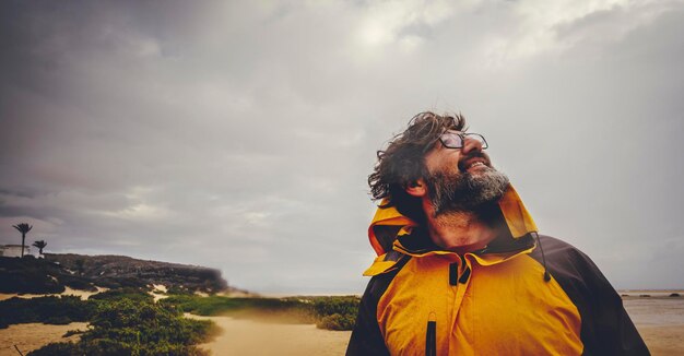 Foto feche de homem maduro, aproveitando o mau tempo e explore a praia ao ar livre. céu nublado e chuvoso. felizes homens livres com jaqueta amarela sorriem e olham para cima