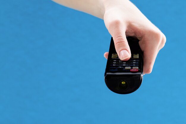 Foto feche de mãos femininas segurando um controle remoto preto para alternar os canais na tv em um fundo azul.