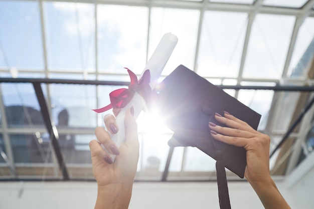 Feche de mãos femininas segurando um diploma e um chapéu de formatura na luz do sol, copie o espaço