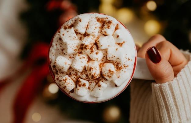 Feche de mãos femininas segurando uma caneca branca com chocolate quente, chá ou café e marshmallow. Conceito de época de inverno e Natal.