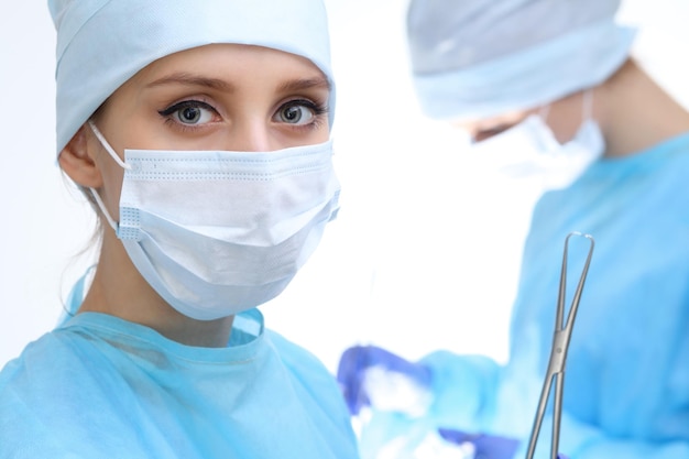 Feche de mulher cirurgiã olhando para a câmera enquanto colegas atuando em segundo plano na sala de operação, isolado. conceito de cirurgia e medicina