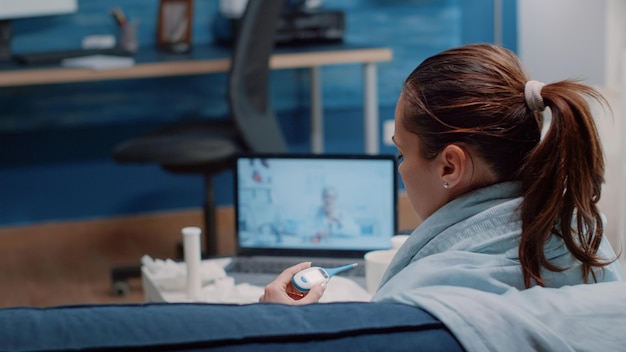 Feche de mulher segurando termômetro com médico em videoconferência. Paciente doente usando telemedicina para curar sintomas de febre e vírus, olhando para instrumento médico para temperatura
