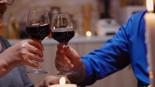 Feche de taças de vinho tinto tilintando durante um jantar romântico. Feliz alegre casal de idosos sênior jantando juntos na cozinha aconchegante, apreciando a refeição, comemorando seu aniversário.