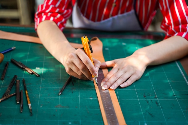 Feche e corte as mãos do fabricante de couro usando uma faca de corte para cortar um pedaço de folha de couro