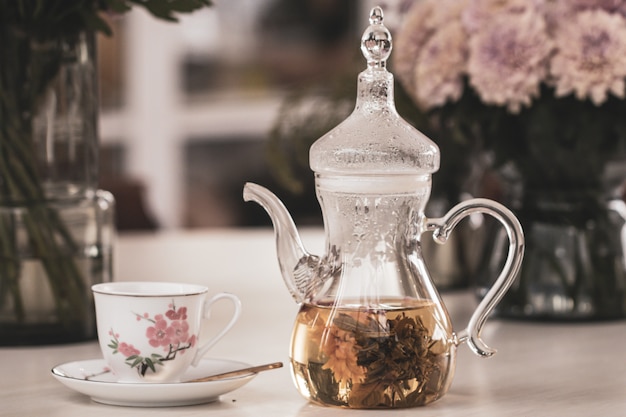Feche o bule e xícara com flor desabrochando chá e flores de crisântemo rosa em vaso de vidro na mesa.