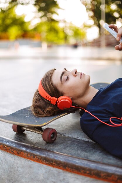 Feche o cara pensativo em fones de ouvido laranja deitado no skate enquanto estiver usando o celular no skatepark